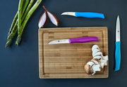 Bovenaanzicht van een driedelige messenset met kleurrijke handgrepen op een snijplank, omringd door asperges, halve uien en gesneden champignons.