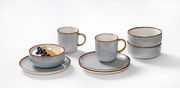 Frühstücksset Portofino 12-tlg in grauer Keramik mit braunem Rand, bestehend aus Tassen, Schalen und Tellern, auf weißem Hintergrund aus seitlicher Perspektive.
