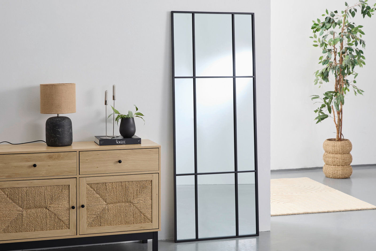 Großer rechteckiger Spiegel mit schwarzem Rahmen, an eine weiße Wand gelehnt, neben einer Holzkommode mit Dekorationen, aus seitlicher Perspektive.