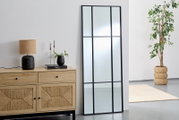 Großer rechteckiger Spiegel mit schwarzem Rahmen, an eine weiße Wand gelehnt, neben einer Holzkommode mit Dekorationen, aus seitlicher Perspektive.