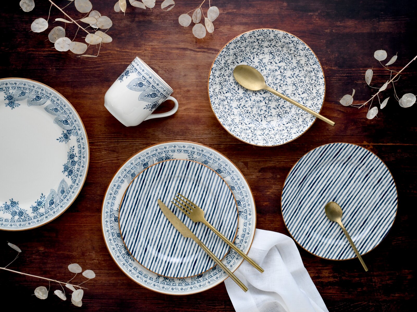 Draufsicht auf blau-weiß gemustertes Geschirr-Set mit Tellern und Tasse sowie goldfarbenem Besteck auf dunklem Holztisch, dekoriert mit weißen Trockenblättern und Stoffserviette