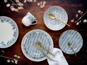Draufsicht auf blau-weiß gemustertes Geschirr-Set mit Tellern und Tasse sowie goldfarbenem Besteck auf dunklem Holztisch, dekoriert mit weißen Trockenblättern und Stoffserviette