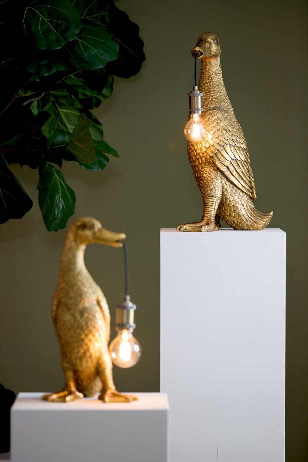 Zwei goldene Tischleuchten in Form von Enten, die Glühbirnen im Schnabel halten, auf weißen Sockeln vor einem grünen Hintergrund. Perspektive von vorne.