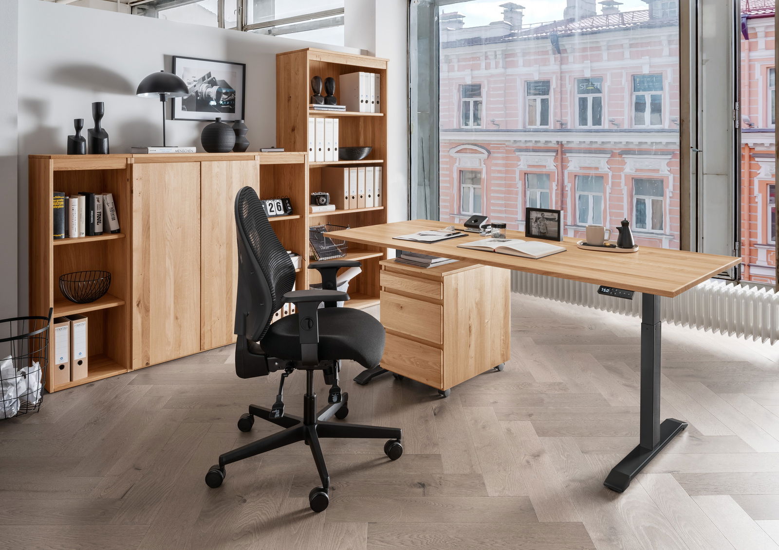 Linea Natura E-bureau DURABLE OFFICE Moderne Büroumgebung mit höhenverstellbarem Schreibtisch, Rollcontainer und ergonomischem Bürostuhl aus der Perspektive eines großen Fensters mit Blick auf ein Gebäude.