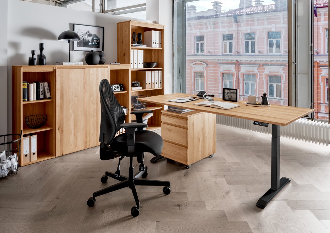 Linea Natura E-Schreibtisch DURABLE OFFICE Moderne Büroumgebung mit höhenverstellbarem Schreibtisch, Rollcontainer und ergonomischem Bürostuhl aus der Perspektive eines großen Fensters mit Blick auf ein Gebäude.