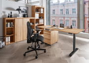 Moderne Büroumgebung mit höhenverstellbarem Schreibtisch, Rollcontainer und ergonomischem Bürostuhl aus der Perspektive eines großen Fensters mit Blick auf ein Gebäude.