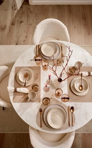Elegantes Tafelservice für vier Personen auf einem runden Marmortisch, aus der Vogelperspektive fotografiert. Enthält Teller, Schalen, Besteck und Gläser in warmen Erdtönen, dekoriert mit Kerzen und Zweigen.