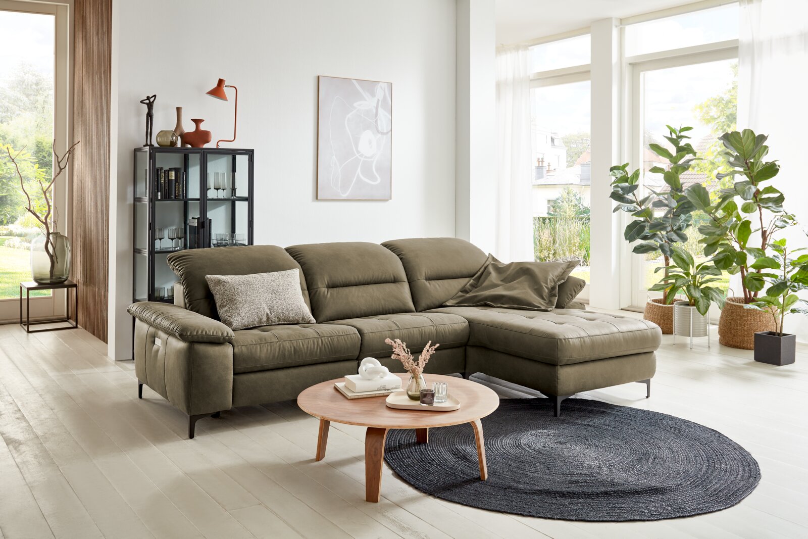 Pure Home Comfort Longchairkombination PURE COMFORT 820 Wohnzimmer mit einer olivgrünen Longchairkombination PURE COMFORT 820, von vorne betrachtet. Die Couch steht in einem hellen Raum mit großen Fenstern, Pflanzen und einem runden Holztisch.
