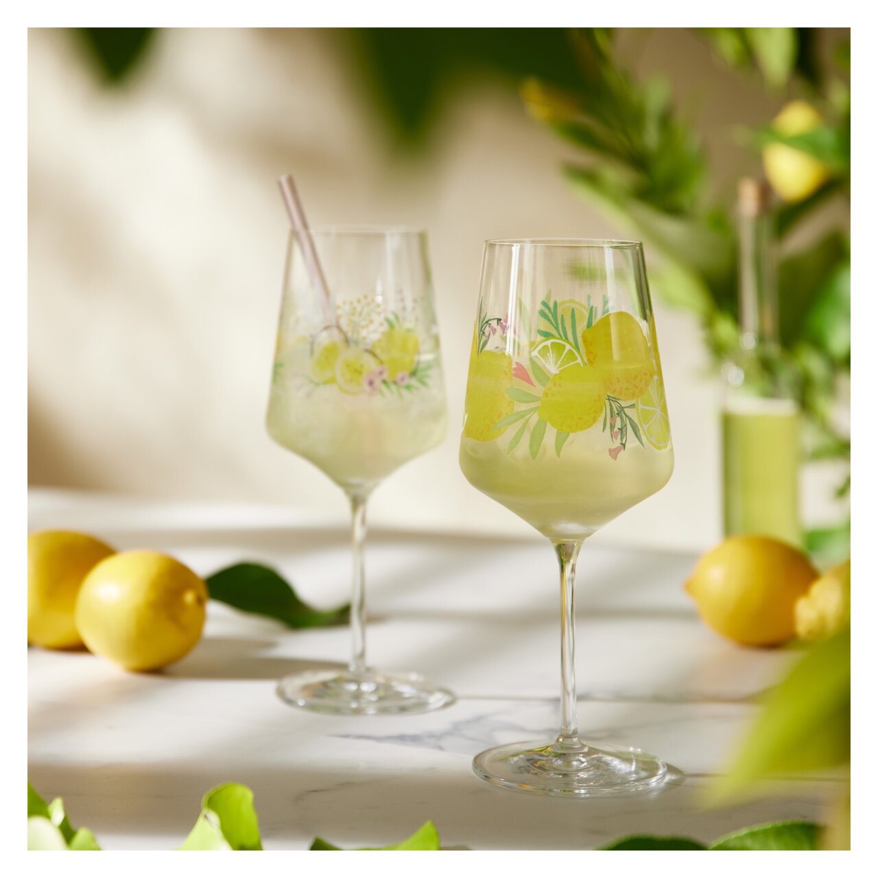 Ritzenhoff Limoncello 2er-Set SOMMERTAU Zwei elegante Gläser mit Limoncello-Motiv, gefüllt mit einem erfrischenden Getränk, umgeben von Zitronen und grünen Blättern, aus einer seitlichen Perspektive fotografiert.