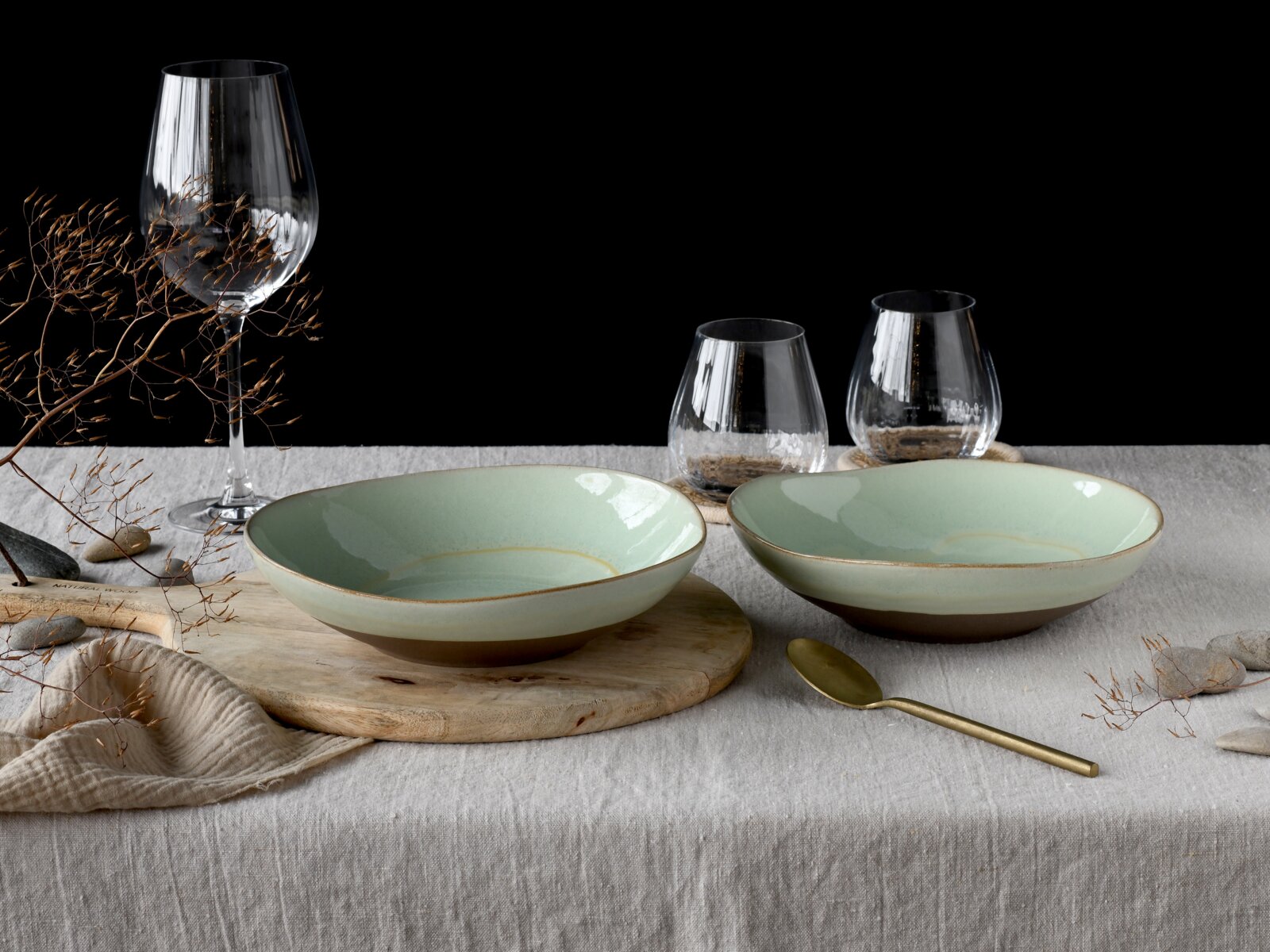 Creatable Pasta Bowl 2-tlg BASALT BIO Zwei flache, mintgrüne Keramikschalen mit braunem Fuß auf Leinentischdecke, daneben goldener Löffel und Glaswaren; frontale Tischansicht mit leicht erhöhter Perspektive vor schwarzem Hintergrund