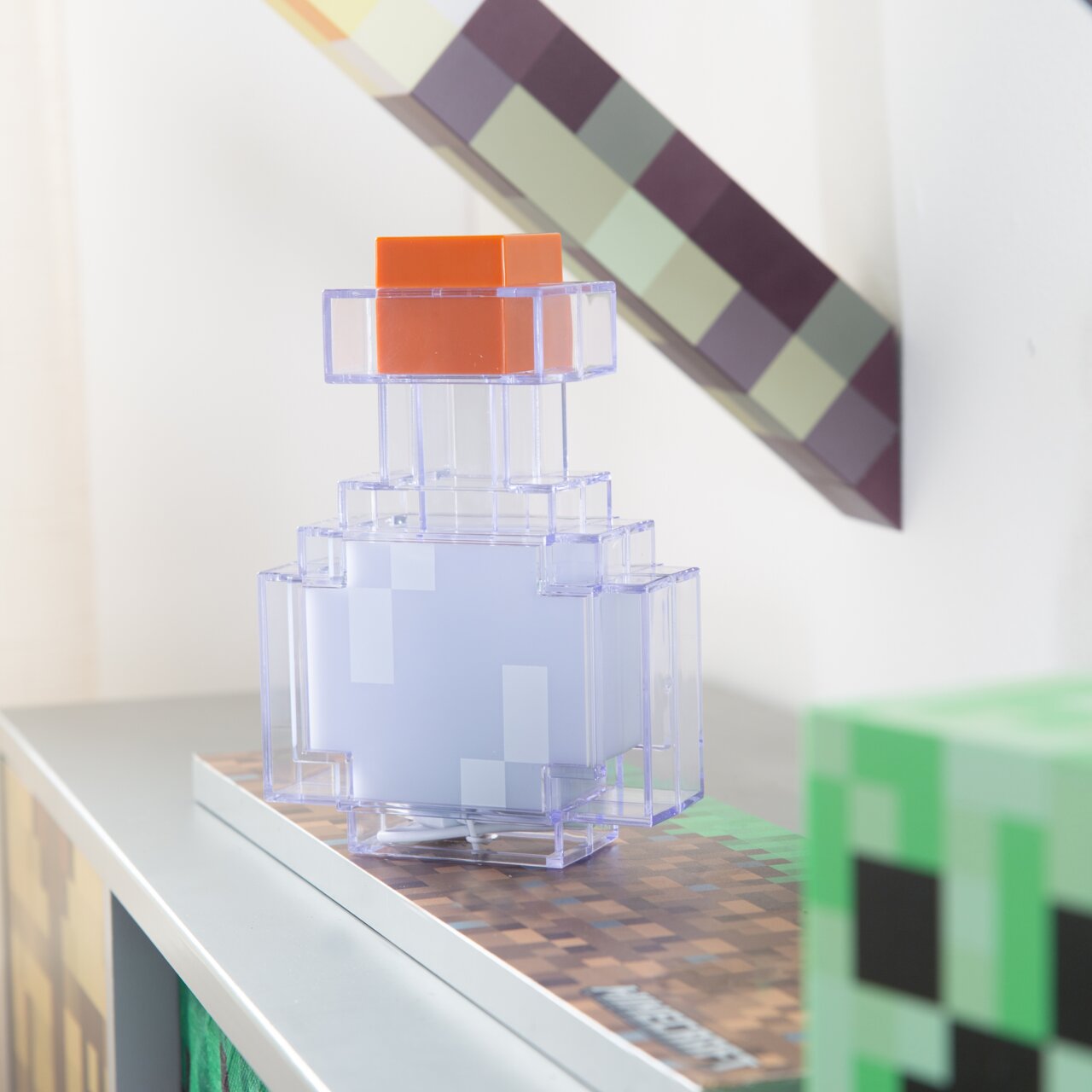 Transparente, pixelige Minecraft-inspirierte Flaschen-Deko mit orangefarbenem Deckel auf einer Box, fotografiert in leichter Seitenansicht (3/4-Perspektive)
