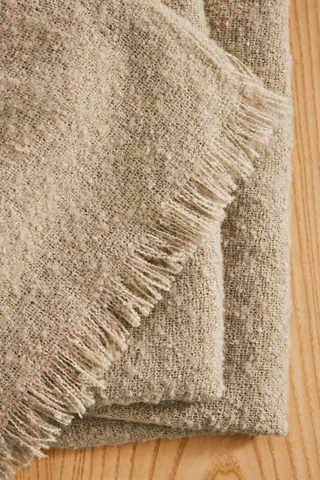 Nahaufnahme einer gefalteten, beigen Wohndecke mit Fransen auf einer Holzoberfläche, von oben betrachtet.