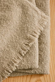 Nahaufnahme einer gefalteten, beigen Wohndecke mit Fransen auf einer Holzoberfläche, von oben betrachtet.