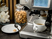 Edelstahl-Kaffeevollautomat/Espressomaschine mit Touchdisplay, die einen Espresso in eine weiße Tasse auf der Abtropfschale brüht; links daneben Keks- und Marshmallow-Gläser sowie Untertasse mit Löffel, aufgenommen aus leicht schräger Frontperspektive von