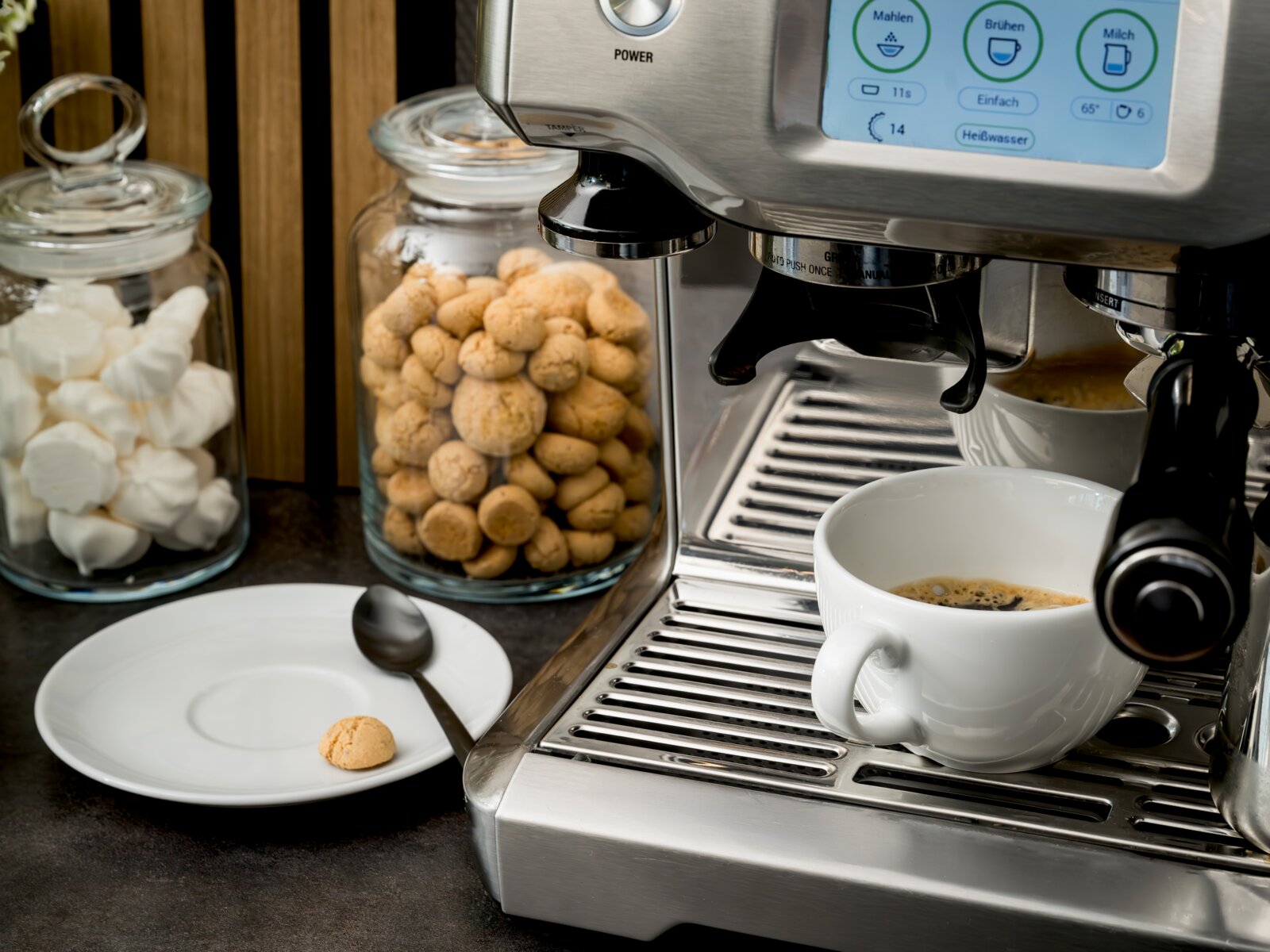 Edelstahl-Kaffeevollautomat/Espressomaschine mit Touchdisplay, die einen Espresso in eine weiße Tasse auf der Abtropfschale brüht; links daneben Keks- und Marshmallow-Gläser sowie Untertasse mit Löffel, aufgenommen aus leicht schräger Frontperspektive von