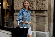 Een vrouw in een blauw geruite blouse draagt een zwarte boodschappenmand in de stad, gezien vanuit een zijaanzicht.