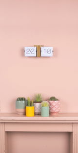 Flip-Uhr ohne Gehäuse in Weiß und Messing, an einer Wand montiert, mit Pflanzen im Vordergrund. Frontalansicht.