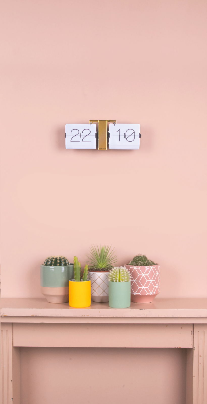 Flip-Uhr ohne Gehäuse in Weiß und Messing, an einer Wand montiert, mit Pflanzen im Vordergrund. Frontalansicht.