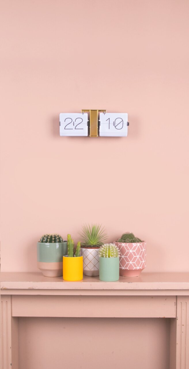 Flip-Uhr ohne Gehäuse in Weiß und Messing, an einer Wand montiert, mit Pflanzen im Vordergrund. Frontalansicht.