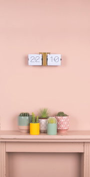 Flip-Uhr ohne Gehäuse in Weiß und Messing, an einer Wand montiert, mit Pflanzen im Vordergrund. Frontalansicht.