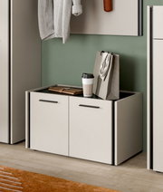 Modernes, niedriges Schuhschrank-/Sideboard-Modul in Weiß mit zwei Türen und schwarzen Griffleisten, oben Ablagefläche mit Kaffeebecher und Tasche, in einer Garderobe vor grüner Wand, perspektivisch in leichter Seitenansicht von vorn fotografiert