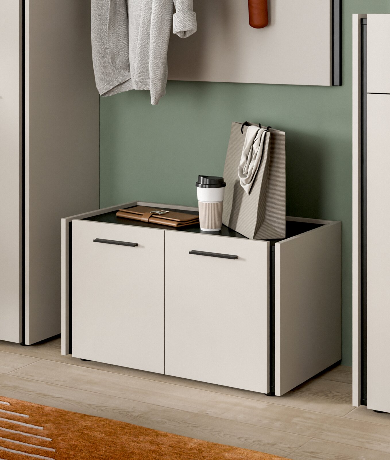 Modernes, niedriges Schuhschrank-/Sideboard-Modul in Weiß mit zwei Türen und schwarzen Griffleisten, oben Ablagefläche mit Kaffeebecher und Tasche, in einer Garderobe vor grüner Wand, perspektivisch in leichter Seitenansicht von vorn fotografiert