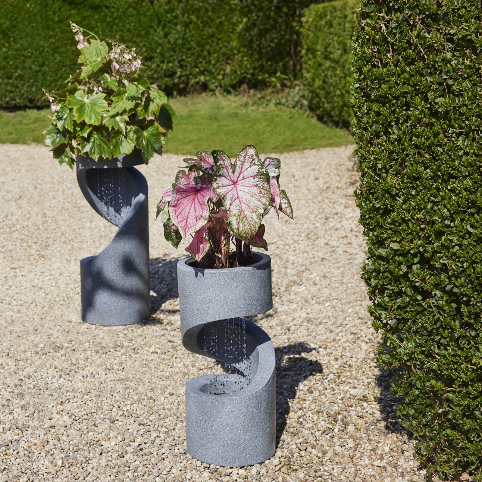 Zwei moderne, gedrehte Pflanzsäulen aus grauem Beton/Steinoptik mit integrierten Pflanzgefäßen und Wasserlauf, in einem Garten auf Kiesweg – frontale Perspektive auf Augenhöhe.
