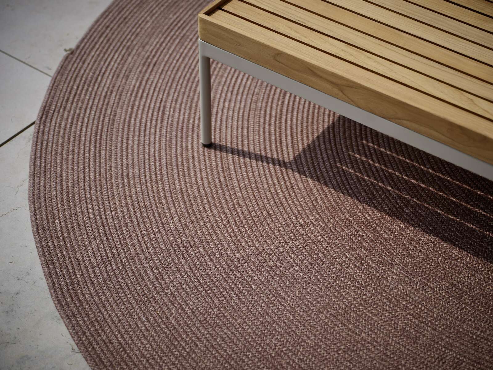 Stern Outdoor Teppich rund Runder Outdoor-Teppich in einem warmen Braunton, teilweise unter einer Holzbank, aus der Vogelperspektive fotografiert.