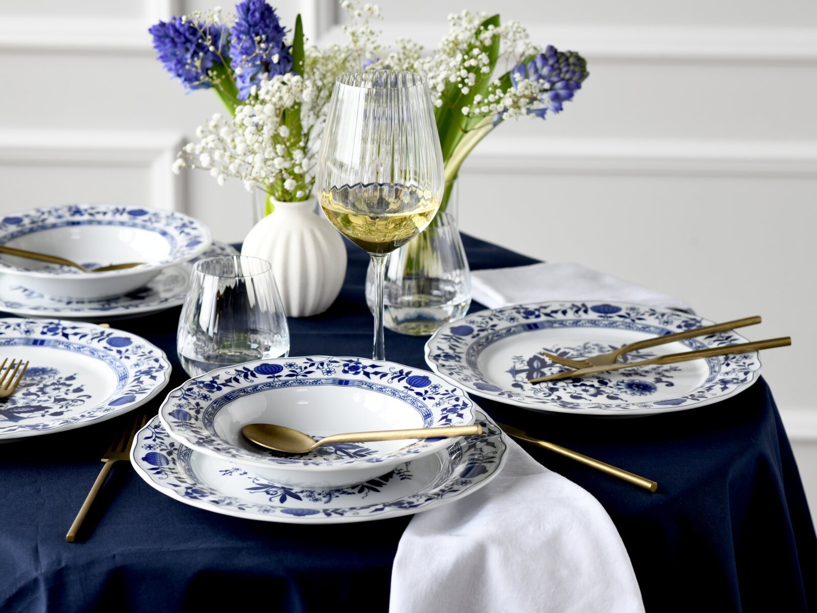 Schick gedeckter Tisch mit blau-weiß gemustertem Porzellangeschirr, goldfarbenem Besteck, gerippten Weingläsern und Blumenvasen mit blauen Hyazinthen und Schleierkraut, aufgenommen in leichter Schrägaufsicht von vorn.