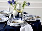 Schick gedeckter Tisch mit blau-weiß gemustertem Porzellangeschirr, goldfarbenem Besteck, gerippten Weingläsern und Blumenvasen mit blauen Hyazinthen und Schleierkraut, aufgenommen in leichter Schrägaufsicht von vorn.