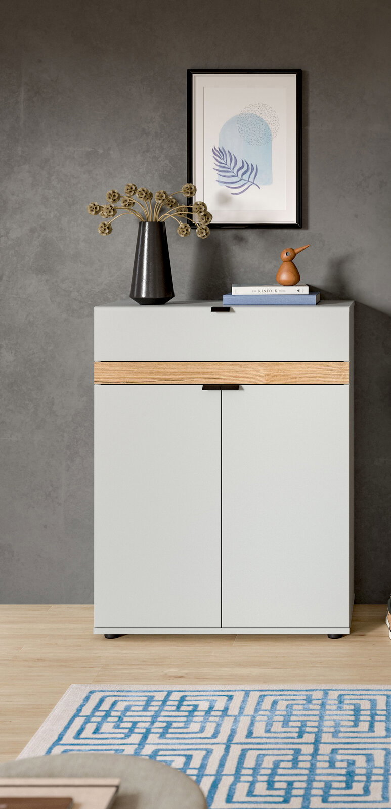 Modernes Sideboard in Hellgrau mit Holz-Akzentleiste und zwei Türen vor grauer Wand, dekoriert mit Vase, Büchern und Holzvogel; Frontalansicht auf Augenhöhe.
