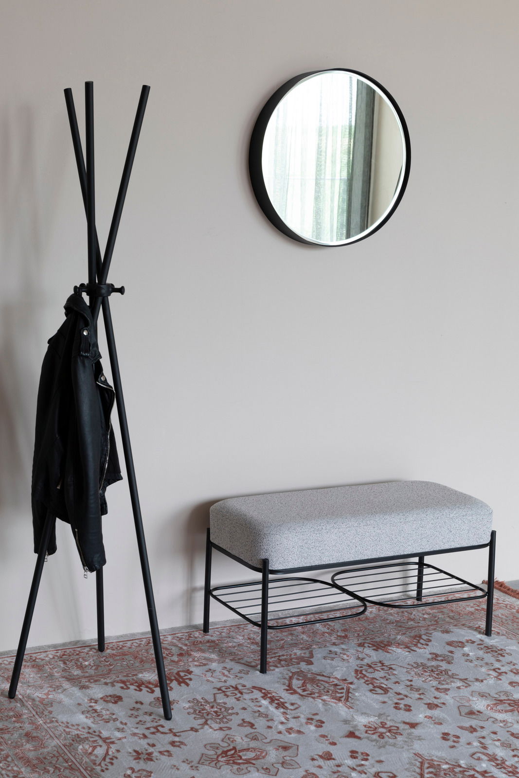 Moderne Sitzbank mit grauem Polster und schwarzem Metallgestell, seitliche Perspektive, neben einem Kleiderständer und einem runden Spiegel an der Wand.
