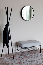 Moderne Sitzbank mit grauem Polster und schwarzem Metallgestell, seitliche Perspektive, neben einem Kleiderständer und einem runden Spiegel an der Wand.