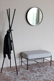 Moderne Sitzbank mit grauem Polster und schwarzem Metallgestell, seitliche Perspektive, neben einem Kleiderständer und einem runden Spiegel an der Wand.