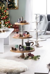 Etagere aus Akazienholz und Aluminium mit drei Ebenen, dekoriert mit Kerzen, Tannenzapfen und einer Kaffeekanne, in einer weihnachtlich geschmückten Wohnzimmerszene, seitliche Perspektive.