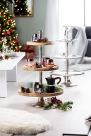 Etagère van acaciahout en aluminium met drie verdiepingen, versierd met kaarsen, dennenappels en een koffiepot, in een kerstachtig versierde woonkamerscène, zijaanzicht.