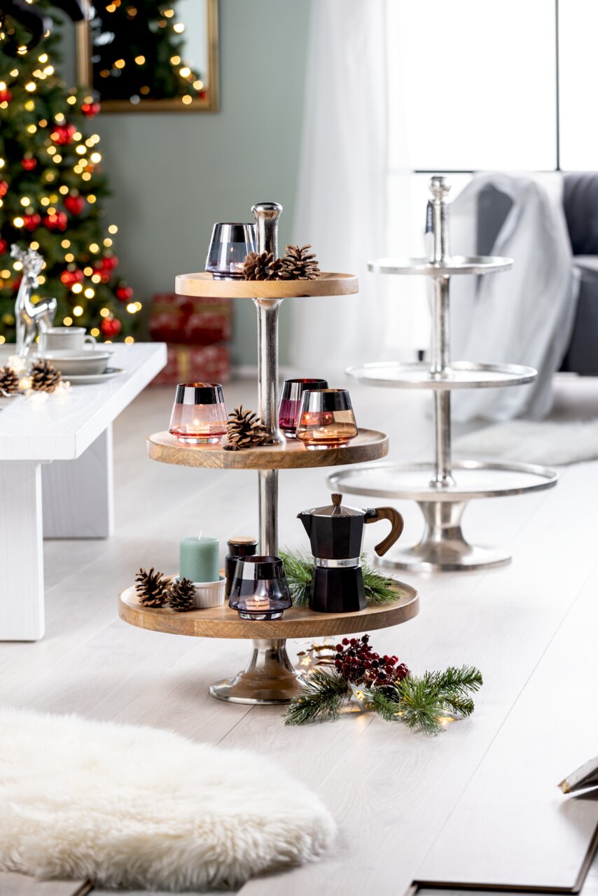 Etagère van acaciahout en aluminium met drie verdiepingen, versierd met kaarsen, dennenappels en een koffiepot, in een kerstachtig versierde woonkamerscène, zijaanzicht.