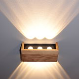Moderne Wandleuchte aus Holz mit drei sichtbaren Lichtquellen, die warmes Licht nach oben und unten an die Wand strahlt. Perspektive von vorne.