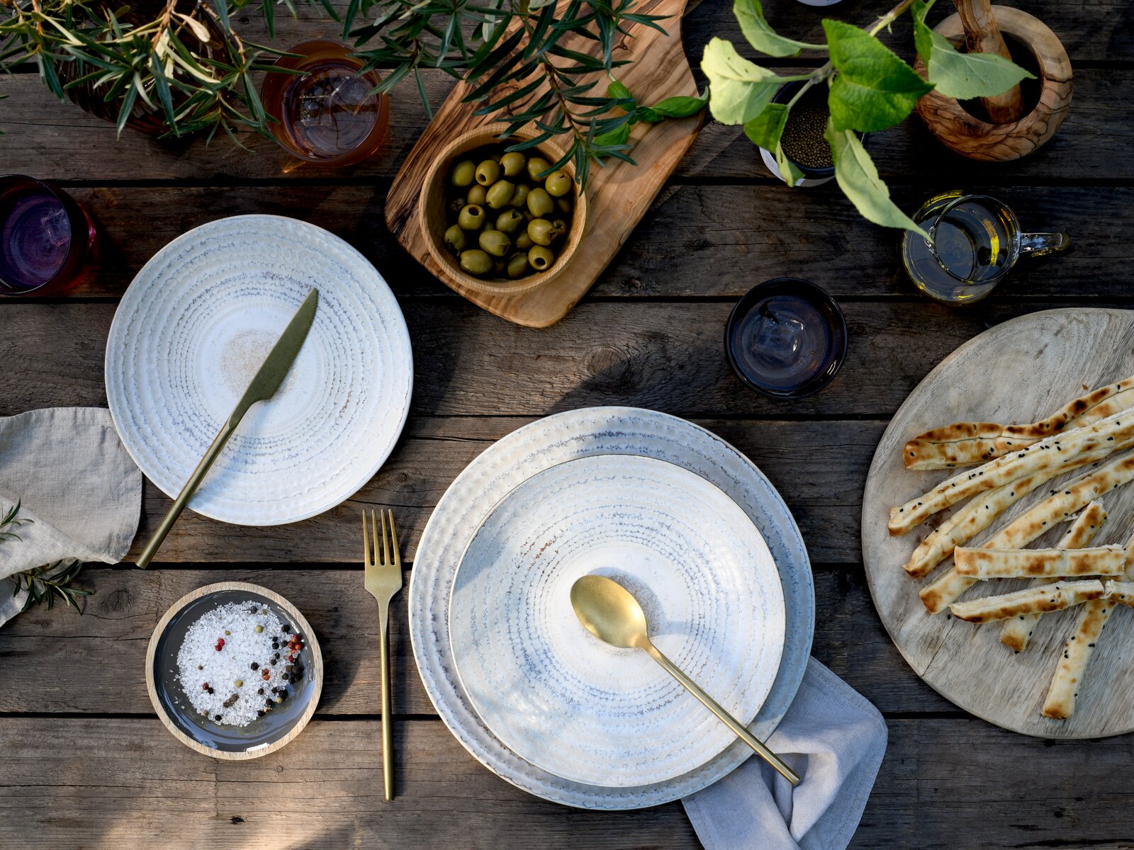 Draufsicht auf rustikal gedeckten Holztisch mit zwei weißen Keramiktellern und goldfarbenem Besteck, Schale grüner Oliven, Salz mit Pfefferkörnern, Brotstangen und Glas Olivenöl zwischen Kräuterzweigen.
