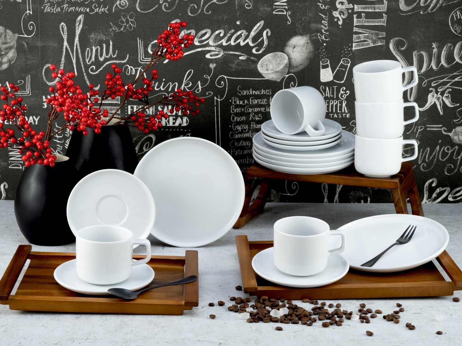 Creatable Kaffeeservice 18-tlg CHEF COLLECTION Schlichtes weißes Porzellan-Geschirrset mit Tassen, Untertassen und Tellern auf Holztabletts und kleinem Holzständer, daneben schwarze Vasen mit roten Beeren vor Tafelwand – Perspektive: leicht erhöhte Frontansicht.