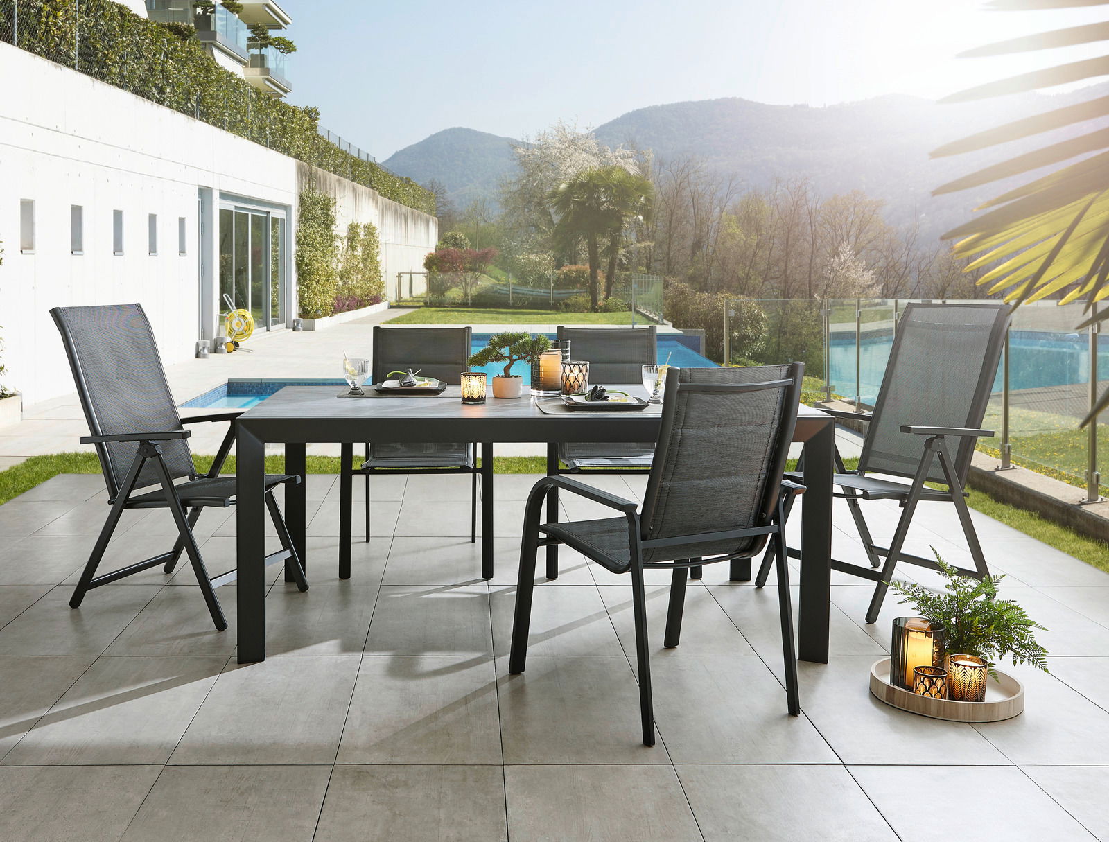 Moderner Gartentisch mit vier Stühlen auf einer Terrasse, umgeben von grüner Landschaft und Pool, aus seitlicher Perspektive fotografiert.