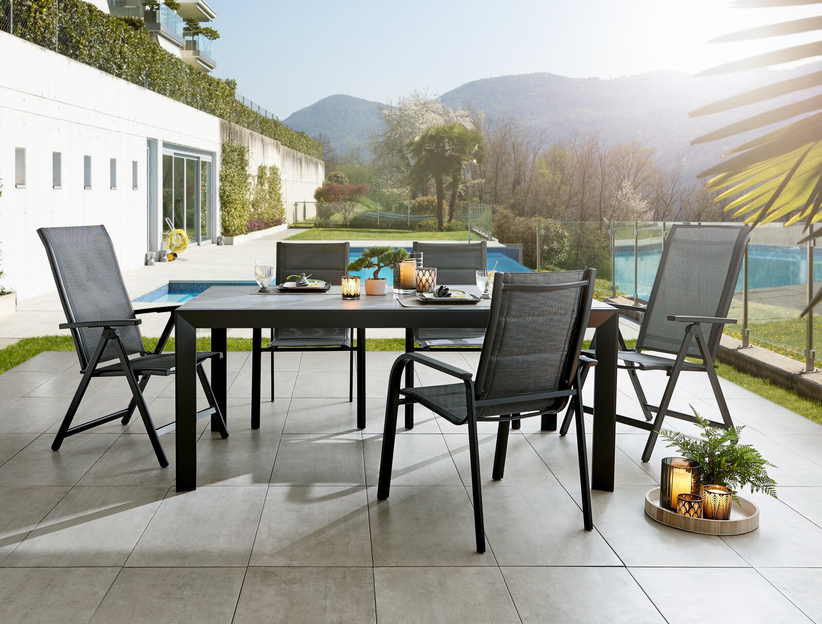Een moderne tuintafel met vier stoelen op een terras, omgeven door een groen landschap en een zwembad, gefotografeerd vanuit een zijdelingse hoek.
