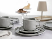Graue Keramik-Kaffeetasse mit eckigem Henkel auf passender Untertasse und Teller, mit schwarzem Löffel; seitliche Nahaufnahme auf Tischgedeck mit weiterer Tasse und Kuchen im Hintergrund