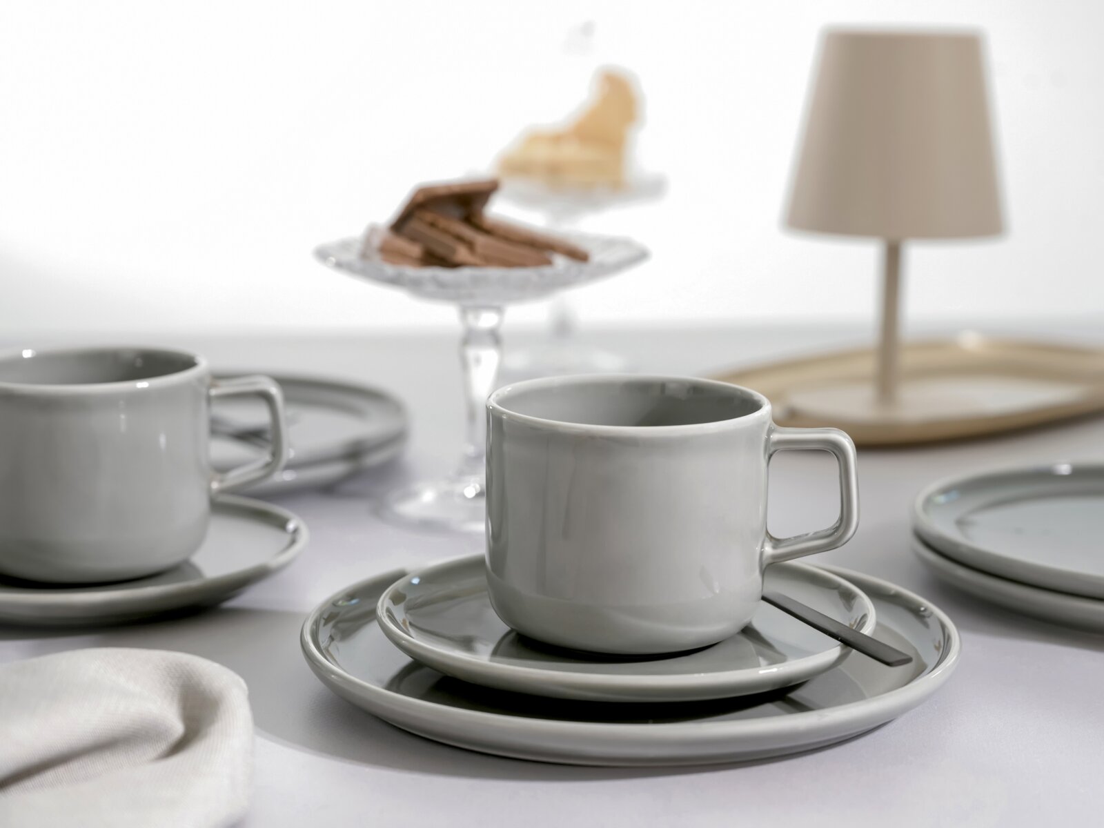 Graue Keramik-Kaffeetasse mit eckigem Henkel auf passender Untertasse und Teller, mit schwarzem Löffel; seitliche Nahaufnahme auf Tischgedeck mit weiterer Tasse und Kuchen im Hintergrund