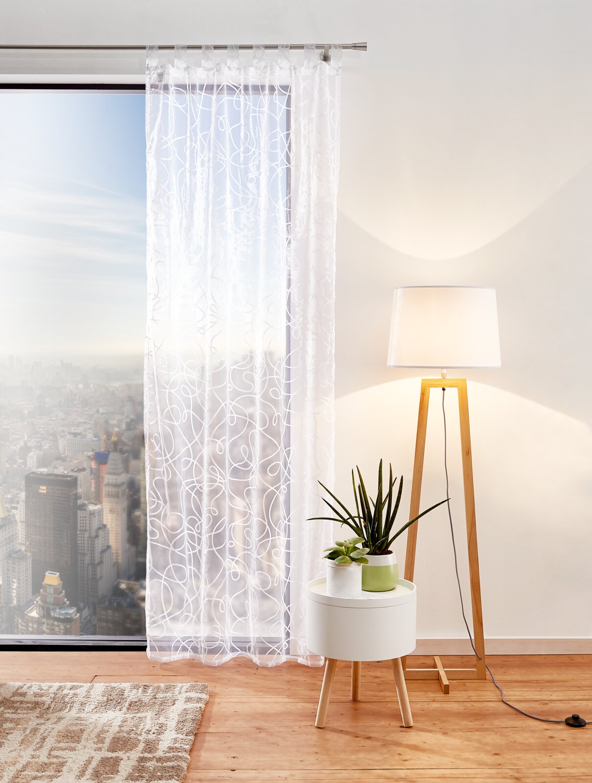 Schlaufenschal Pro 140x245 cm in einem modernen Wohnzimmer, mit Blick auf die Stadt durch ein großes Fenster, neben einem weißen Beistelltisch mit Pflanzen und einer Stehlampe.