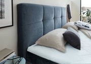 Seitliche Perspektive auf ein Boxbett mit gepolstertem, dunkelblauem Kopfteil und mehreren Kissen in einem modernen Schlafzimmer.