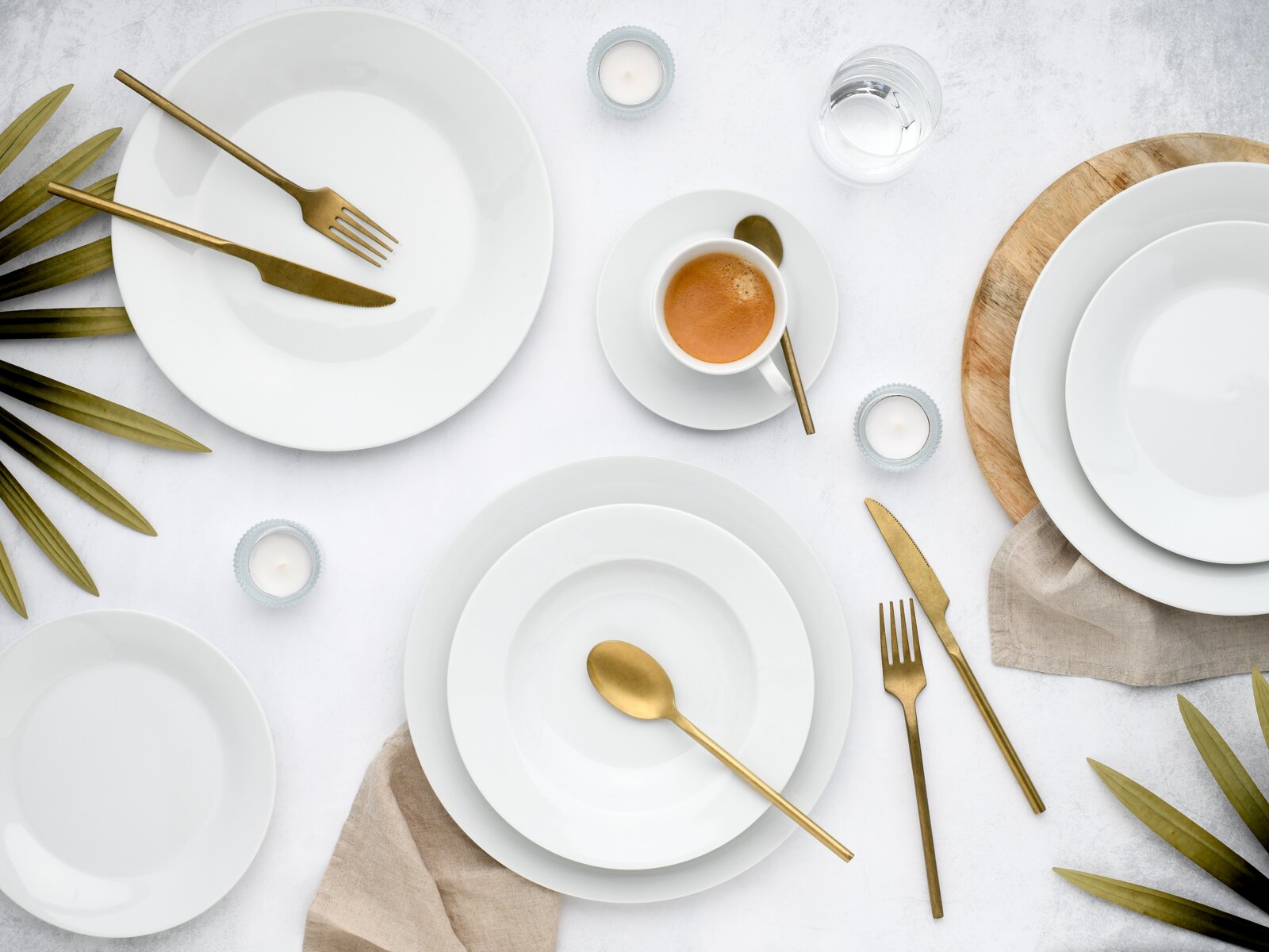 Minimalistisches Tisch-Arrangement mit weißen Porzellantellern, goldfarbenem Besteck und Espresso in Tasse auf Untertasse, ergänzt durch Teelichter, Glas Wasser und grüne Blätter auf hellem Untergrund, Draufsicht (von oben)