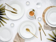 Minimalistisches Tisch-Arrangement mit weißen Porzellantellern, goldfarbenem Besteck und Espresso in Tasse auf Untertasse, ergänzt durch Teelichter, Glas Wasser und grüne Blätter auf hellem Untergrund, Draufsicht (von oben)