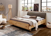 Modernes Holzbett aus Massivholz mit gepolstertem grauem Kopfteil in einem hellen Schlafzimmer, aufgenommen in schräger Frontansicht von links.