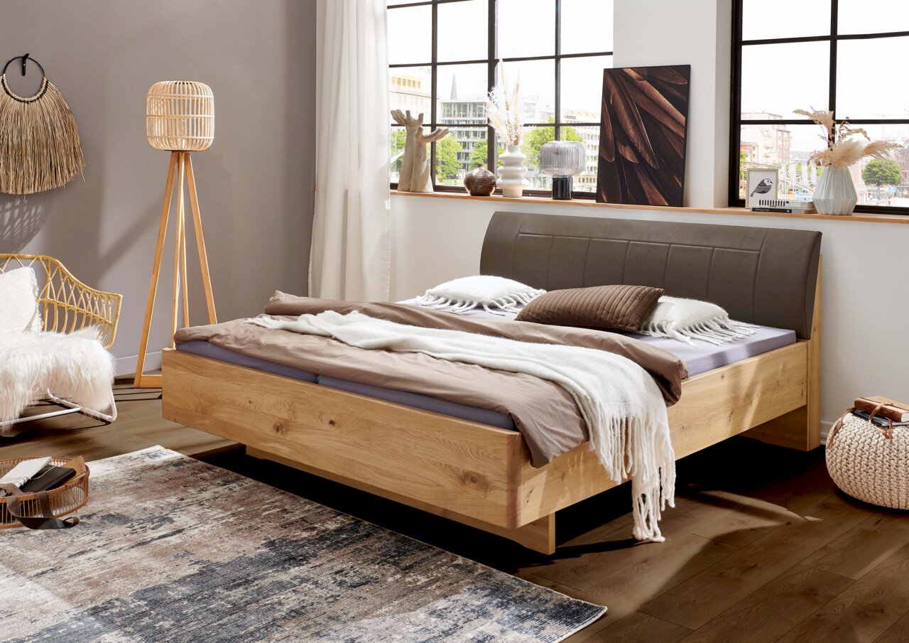 Modern houten bed van massief hout met een gestoffeerd grijs hoofdeinde in een lichte slaapkamer, gefotografeerd vanuit een schuin vooraanzicht vanaf links.